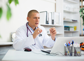 Focused doctor sitting at table in medical office, talking to webcam on laptop, conducting online...