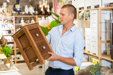 Fototapeta premium Man choosing wooden small cabinet in store. He's standing and holding it in hands.