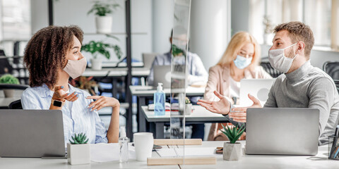 Naklejka premium Meeting during work. Guy and african american woman in protective masks talking about marketing through glass board at workplace with laptops and antiseptic in interior of office during COVID-19