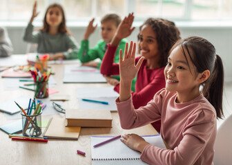Happy Multiethnic School Kids Learning Raising Arms During Lesson Sitting At Desk In Classroom At School. Modern Education And Knowledge Concept. Selective Focus