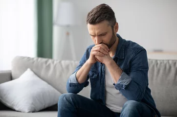 Zelfklevend Fotobehang Upset bearded middle-aged man in casual outfit sitting on sofa in living room, thinking about something, looking down, frustrated man having financial problems during pandemic, copy space © Prostock-studio