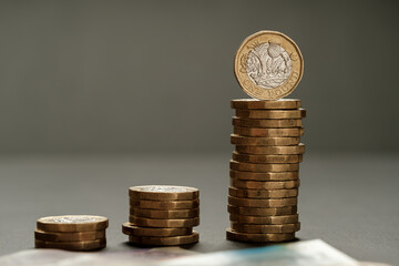 Three increasing stacks of one pound coins arranged in ascending order with blurred banknotes in foreground, illustrating income growth and financial planning, Stafford, United Kingdom, May 18, 2025..