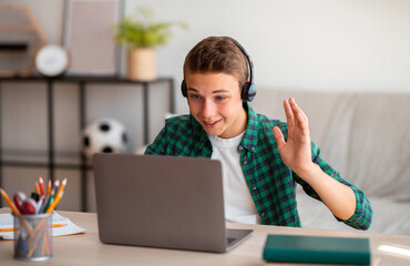 Positive teenager having video conference on laptop with teacher from home, wearing headset and...