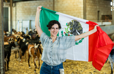 Happy young Latin female traveler waving Mexican flag inside of livestock goat farmhouse