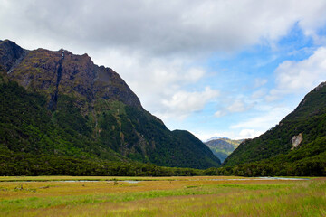 Serene valley framed by lush green mountains under a partly cloudy sky, with a grassy plain leading to a winding river, embodying the tranquility and majesty of untouched nature.