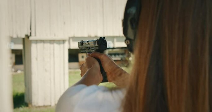 Woman with electronic earmuffs aiming and firing pistol at shooting range targets