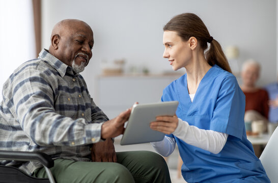 Friendly young lady in workwear nurse helping black senior man in weheelchair using modern digital tablet, elderly african american male patient surfing on Internet at nursing home