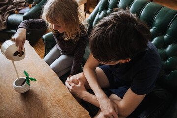 Child watering a small plant while an older sibling observes in a cozy indoor setting
