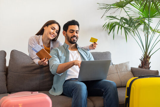 Happy young couple using laptop and booking hotel online with credit card, woman holding passports with tickets, sitting with packed suitcases on sofa at home