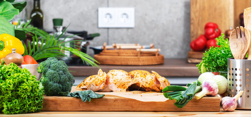 Food background. Healthy eating, cooking and blogging banner. Cutting board with cut raw chicken in marinade, vegetables, fresh herbs on cozy kitchen with wooden table, kitchenware and utensils