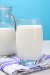 Fresh milk in glass and jug on white wooden table against light blue background, closeup