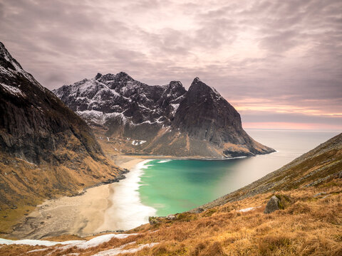 Dramatic high-angle view of the remote Kvalvika Beach on Lofoten Islands, Norway, showcasing the turquoise Arctic ocean, towering snow-dusted mountains, and rugged coastline under a breathtaking paste