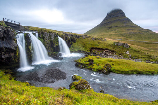 Iconic panoramic view of Kirkjufellsfoss waterfall and the distinct, arrow-shaped Kirkjufell mountain (Church Mountain) in Snaefellsnes Peninsula, Iceland, captured under a dramatic cloudy sky with a 