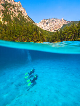 Split-level view of two scuba divers swimming over the clear, sandy bottom of Green Lake (Gr&uuml;ner See) in Styria, Austria, showcasing the phenomenal turquoise water clarity and the dramatic surrounding