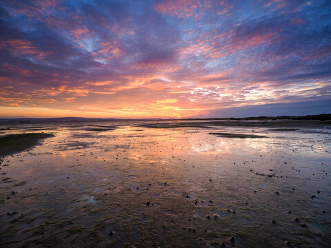 Colorful sunset sky reflected on wet sand during low tide at the beach, creating a tranquil coastal scene at Holy Island at low tide