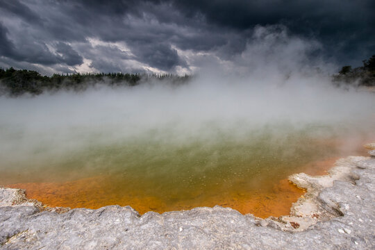 Vibrant Champagne Pool geothermal spring with orange sinter ledge and thick steam under stormy sky, Wai-O-Tapu, New Zealand