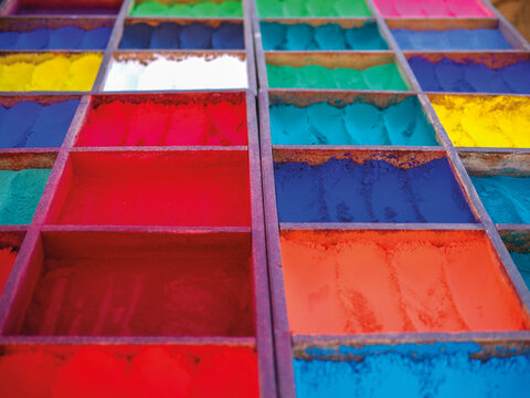 Vibrant powdered pigments arranged in wooden compartments at an outdoor market, viewed from above.