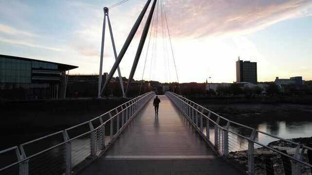 Man walking on modern footbridge over river at dusk. Forwards tracking of person on walkway. Newport, Wales, UK