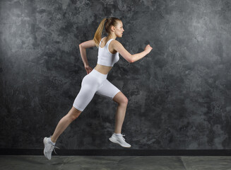 Athletic woman in white sportswear running in motion against dark textured wall. Concept of strength, speed, fitness and active lifestyle. Studio action shot.