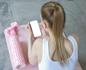 Woman lying on yoga mat after workout, holding smartphone with blank screen. Pink dumbbells and foam roller nearby. Concept of fitness tracking and mobile app use.