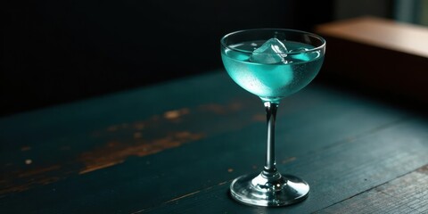 A refreshing teal cocktail in a coupe glass, ice cube melting, sits on a dark wooden surface