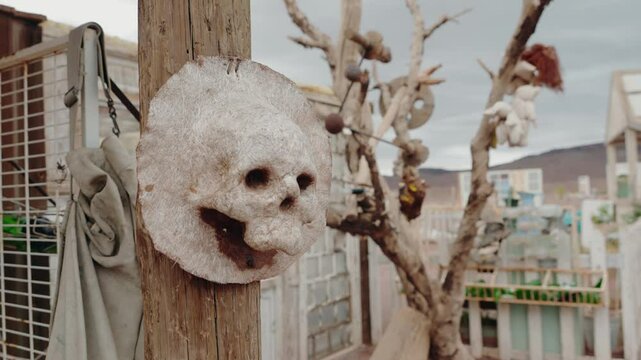 Smiling skull mask hangs on a wooden post in a deserted area with other strange objects and a creepy doll hanging from a dead tree in the background. Eerie and unsettling scene.