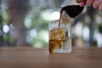 Hand pouring americano in glass with ice cubes and cool water on wood table