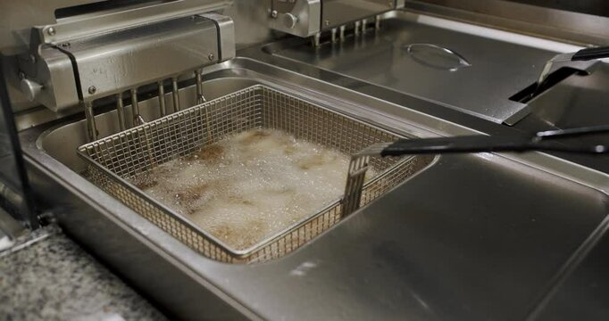 Close-up of french fries cooking in a deep fryer in a restaurant kitchen. The fries are bubbling in hot oil as tongs lift the metal basket. The stainless steel deep fryer reflects the light.