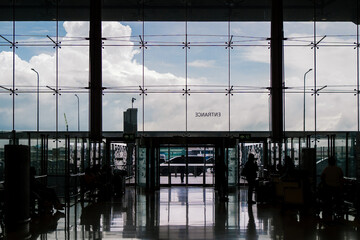 Silhouettes of travelers waiting near a large glass entrance at an airport, with clouds and sky...