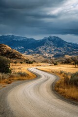 Fototapeta premium Winding Gravel Road Leading to Distant Snowy Mountains under Dramatic Sky in Rural Landscape View