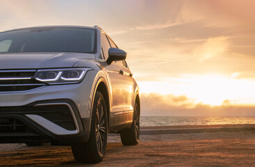 A close-up of a gray car parked on the seashore at sunset. The soft light highlights its silhouette, creating a sense of solitude and freedom against the calm waves and vibrant sky.