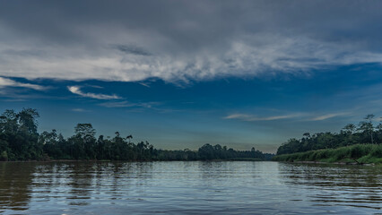 A calm tropical river in the jungle. Early morning. Thickets of rain forest on the shores. Blue sky, clouds. Reflection on the shiny water. Malaysia. Borneo. Sandakan.Kinabatangan River Nature Reserve