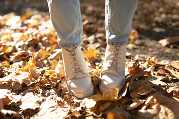 Female legs in light jeans and white leather boots. Woman walking in autumn park during daytime with fallen leaves