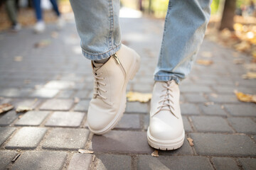 Female legs in light jeans and white leather boots. Woman walking in autumn park during daytime with fallen leaves