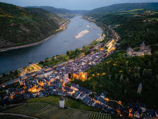 Aerial drone view of Bacharach, Germany, at dusk. The historic town in the UNESCO Upper Middle Rhine Valley glows with evening lights, featuring illuminated Stahleck Castle and churches by the Rhine.