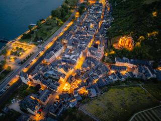 Aerial drone view of Bacharach, Germany, at dusk. The historic town in the UNESCO Upper Middle Rhine Valley glows with evening lights, featuring illuminated Stahleck Castle and churches by the Rhine.