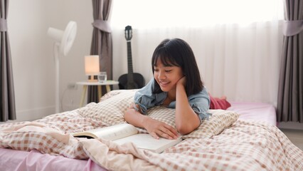 Asian girl smiling while reading book and studying at home in cozy bedroom, representing relaxed...