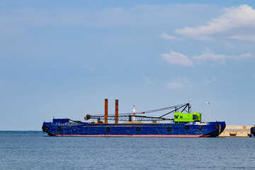 A vibrant blue ship, equipped with cranes and various tools, is anchored close to the rocky coastline nearby