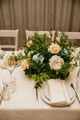 On a table covered with a white tablecloth stands white dishes served for a holiday or banquet, napkins lie on the plates, glasses are nearby and a bouquet of fresh flowers in the center
