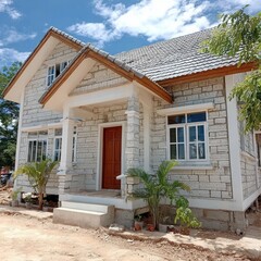 White stone house with brown wooden door and roof, under blue sky with some clouds. Small plants are visible near the house's entrance. The house is a single story structure with multiple windows.