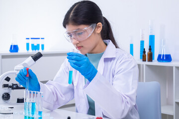 A young Asian female scientist in lab coat and gloves holds a glass pipette carefully in a science...