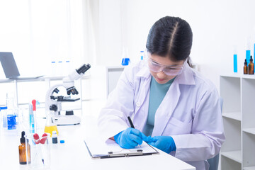 Asian female science student in lab coat writes notes in a laboratory notebook while analyzing experiment, engaging in science, laboratory, scientist, experiment, and research documentation.