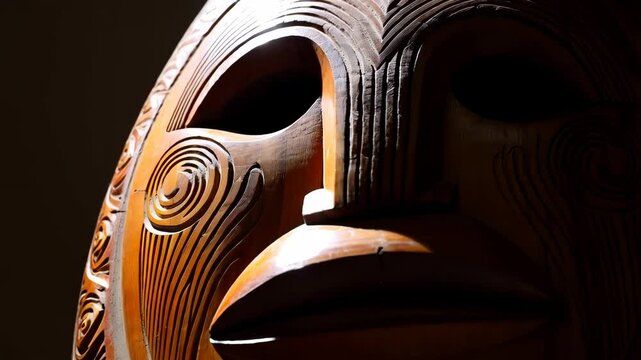 Dramatic close-up of a carved wooden face mask with detailed swirling patterns and a rich brown tone, illuminated by a light source.