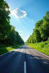 Fototapeta premium A sunlit asphalt road stretches ahead, bordered by lush green trees casting shadows on the pavement, a picturesque summer scene.