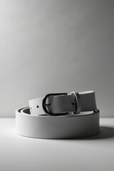 A simple, elegant off-white leather belt with a subtly contrasting dark grey buckle, neatly coiled on a smooth surface, creates a minimalist fashion accessory image.