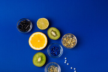 Many vitamins and minerals, dietary supplements in small glass bowls, orange, kiwi, lime and lemon as sources of natural vitaminsa nd minerals. Vitamins and fruits from above on a blue background