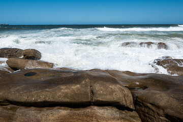 Powerful ocean waves crash against rugged coastal rocks under a clear blue sky. This dynamic seascape captures the raw energy of the ocean and the texture of weathered sandstone, making it ideal for t
