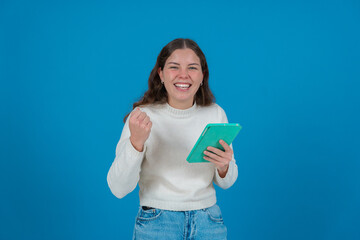 Happy young woman in a white sweater raising her fist in celebration while holding a green tablet and smiling at the camera, against a blue background, symbolizing success, joy, and digital life. 