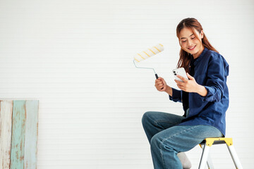 Young woman sitting on a ladder holding a paint roller and smartphone, smiling while planning her...