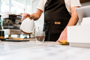 Barista in apron pours hot water into a glass mug for fresh drip coffee at a modern cafe counter. Close-up scene ideal for themes on beverages, barista work, small business, and cafe culture.